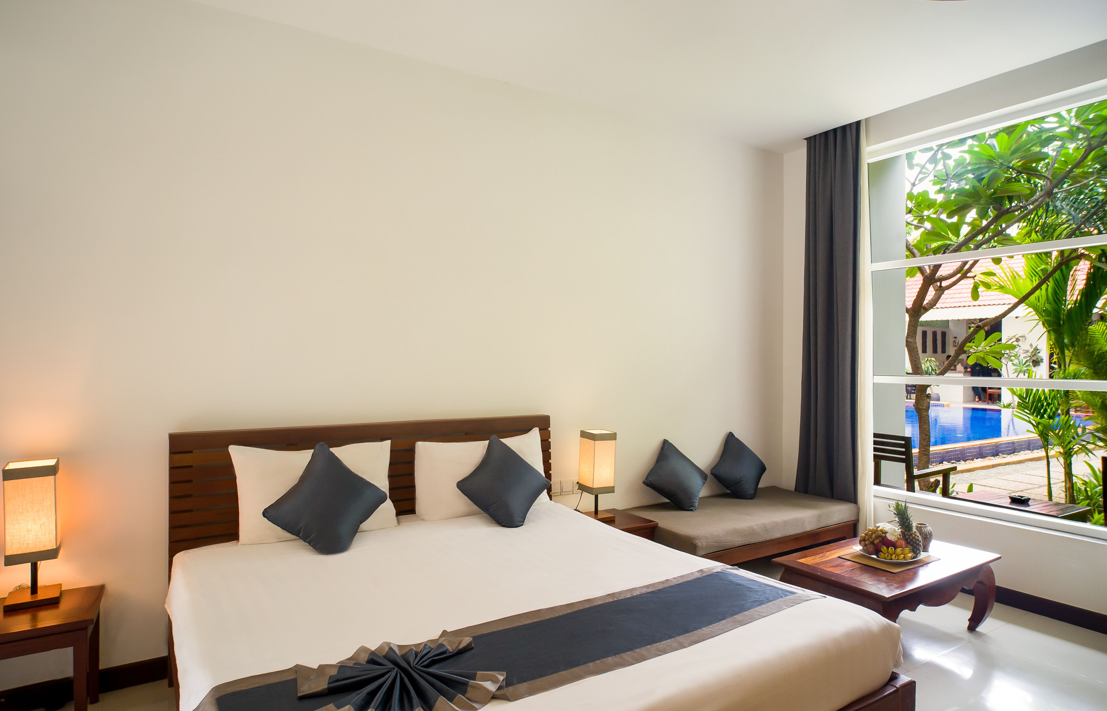 Ein Hotelzimmer mit einem großen Bett, einer Sitzecke, einem kleinen Tisch mit Obst und Blick auf einen Pool und Grünflächen.