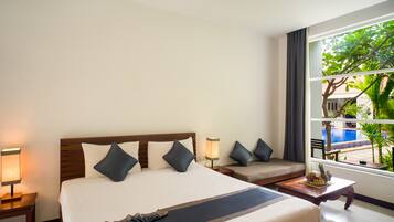 Ein Hotelzimmer mit einem großen Bett, einer Sitzecke, einem kleinen Tisch mit Obst und Blick auf einen Pool und Grünflächen.