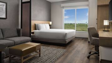 A modern hotel room with a bed, a sofa, a desk, and a large window offering a view of the outdoors.