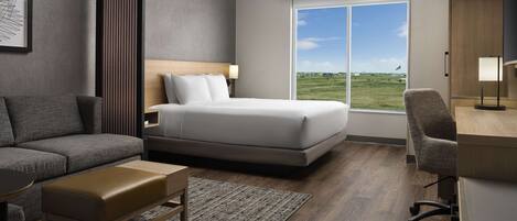 A modern hotel room with a bed, a sofa, a desk, and a large window offering a view of the outdoors.