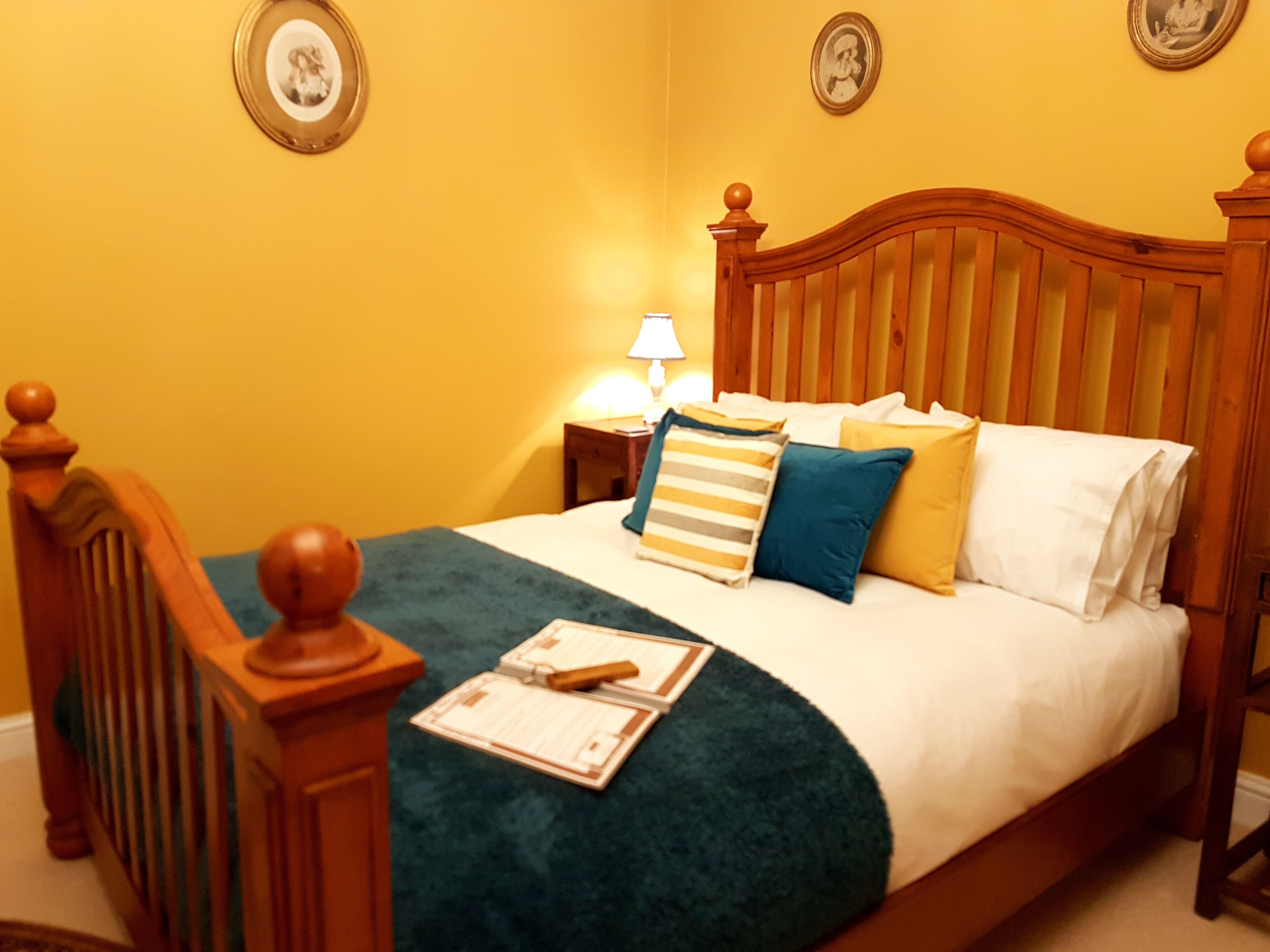 A bed with a wooden headboard, a green throw blanket, and multiple pillows in a room with yellow walls and framed pictures on the wall.
