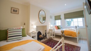 A bedroom with two beds, a bedside table with a lamp, a mirror, and a window with a view. - The Portland Guesthouse (Cheltenham)