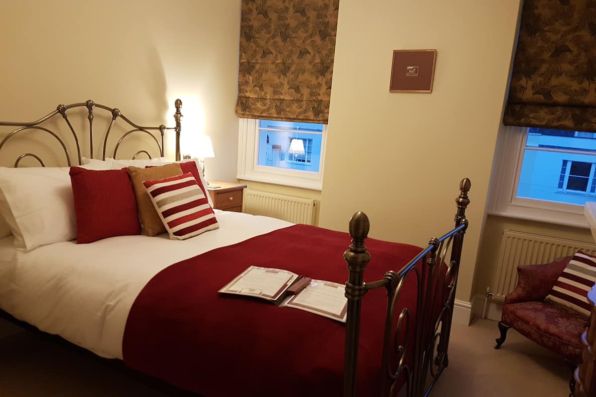 A neatly made bed with a red and white striped pillow, a wooden headboard, and a red throw blanket. There is a small bedside table with a lamp and a framed picture on the wall.