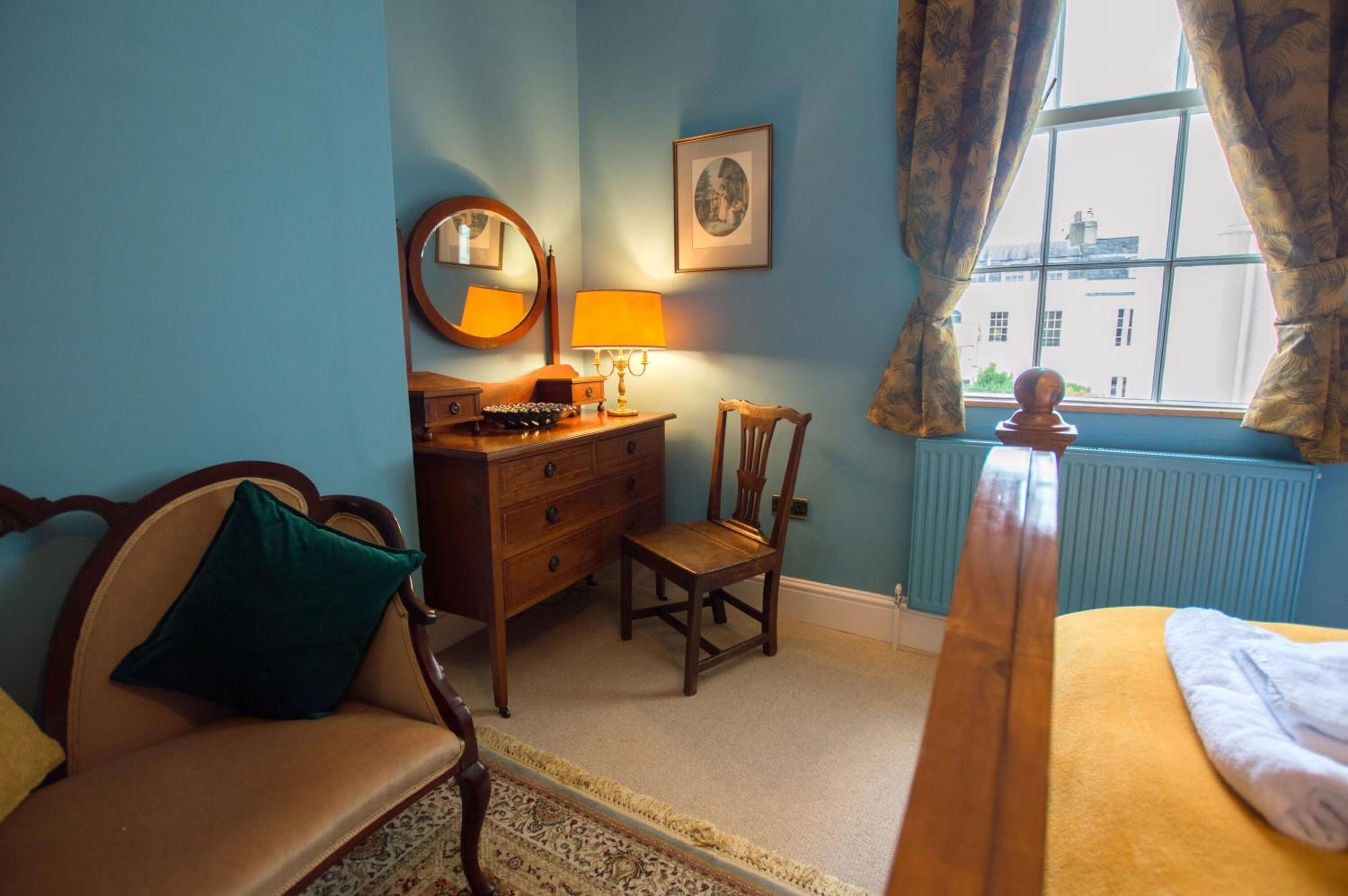 Een kamer met een houten dressoir, een stoel, een bed, een raam met gordijnen, een spiegel en een lijst met een afbeelding aan de muur.
