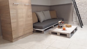 A cozy corner with a wooden cabinet, a grey sofa with striped and solid cushions, and a small wooden table with a candle and a fruit bowl. - Central Cozy Cute Downtown Studio 1BR (Athens)