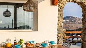 Una mesa lista para el desayuno, con vistas a un paisaje montañoso a través de un arco, y lámparas de mimbre colgantes.