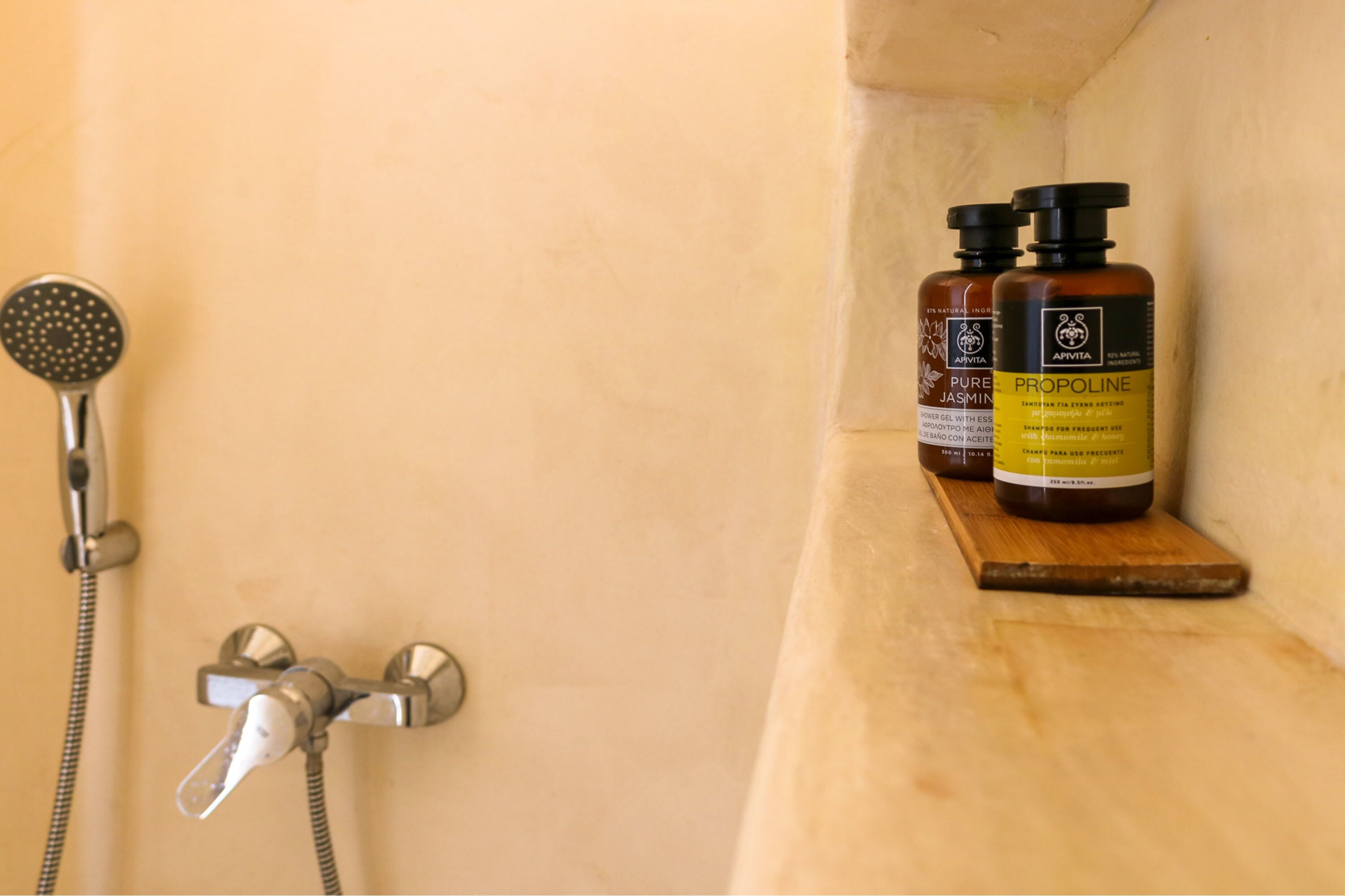 A shower area with a handheld showerhead and a wall-mounted faucet. Two bottles of Apivia Pure Jasmine and Propylene are placed on a wooden shelf.