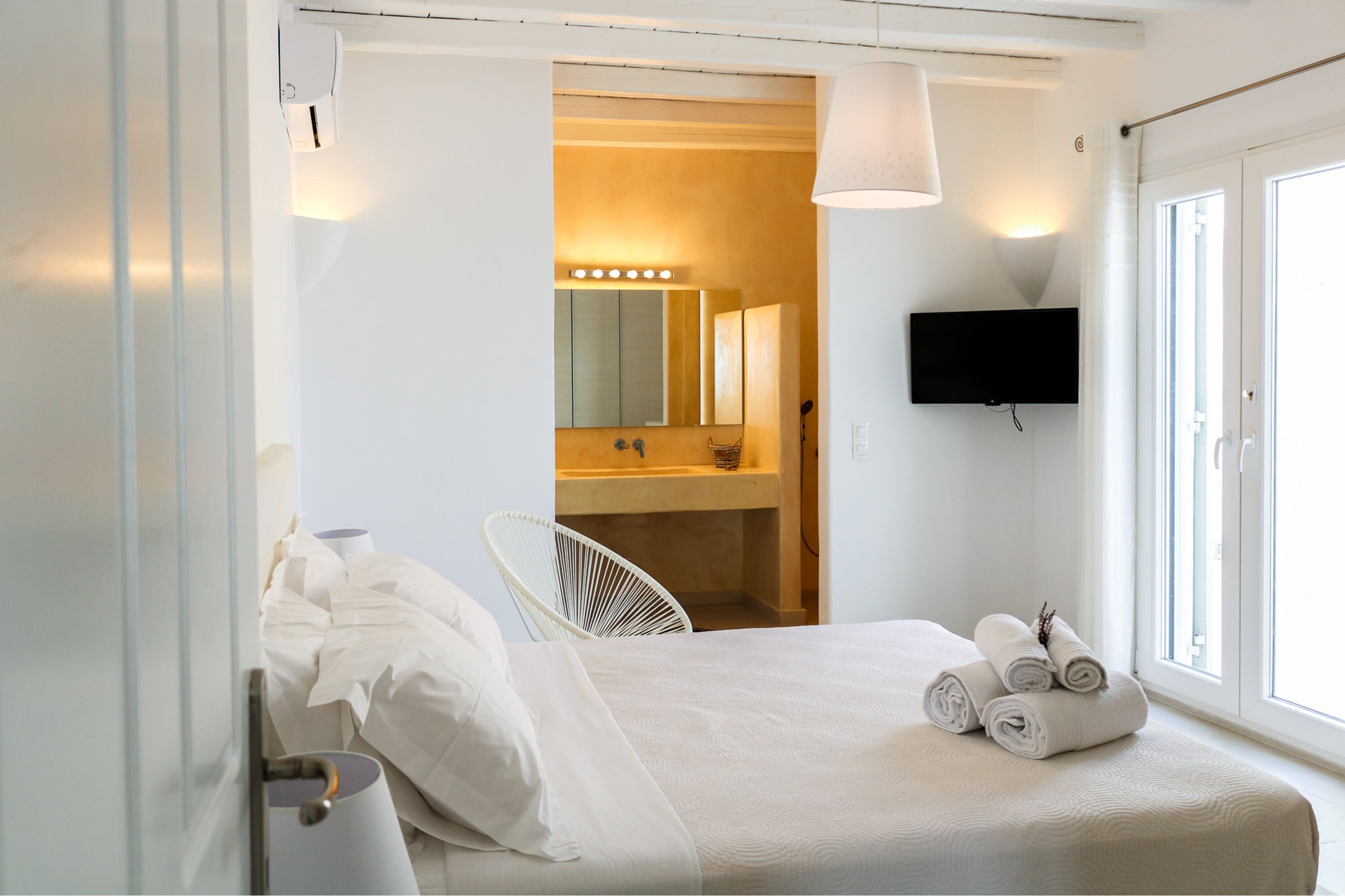 Habitación de hotel con cama, una silla, televisor, baño con espejo e iluminación, y ventana.