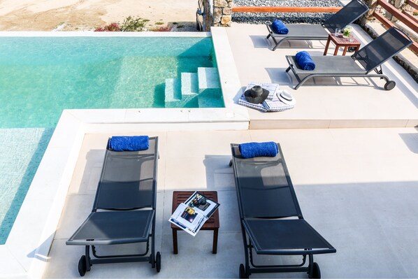 Zona junto a la piscina con dos tumbonas, una mesita con una revista, y una pared de piedra al fondo.