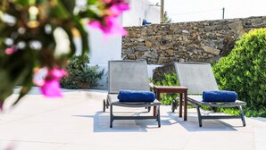 Un espacio al aire libre iluminado por el sol con dos sillones de relax, cada uno con un cojín azul, situados frente a una pared de piedra y rodeados de follaje.