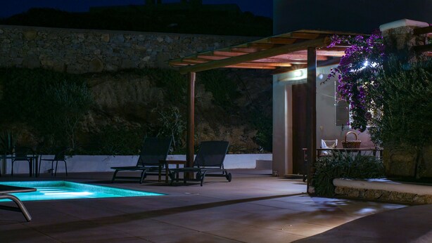 Un área de piscina al aire libre con dos sillas de descanso, una pérgola y un muro de piedra.