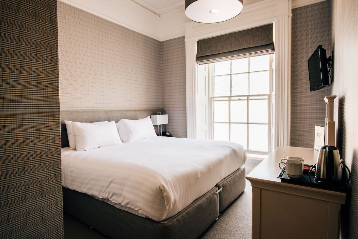 A neatly made bed with a headboard, a bedside table with a lamp and a cup, and a window with blinds.