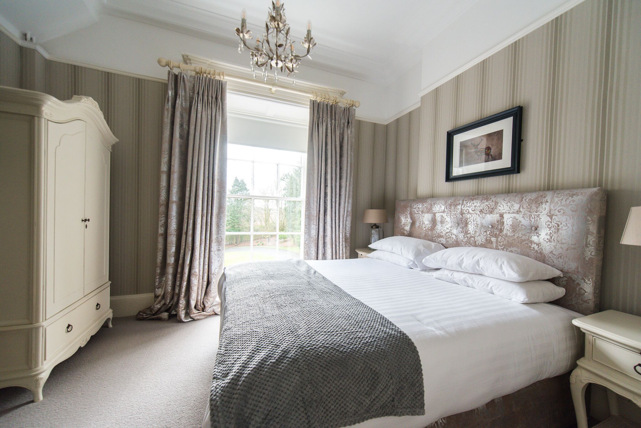 A bedroom with a large bed, a wardrobe, a chandelier, and a window with curtains.