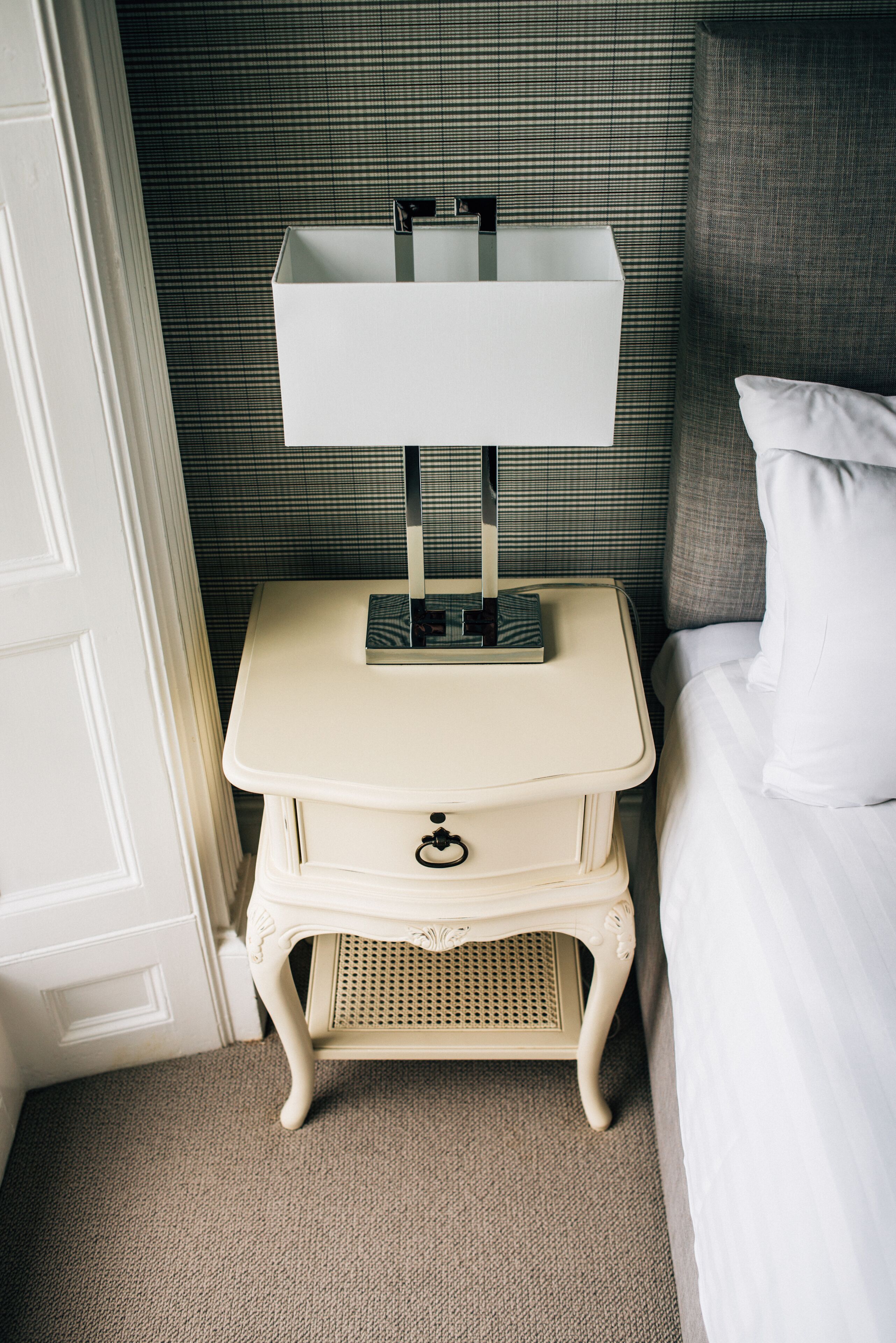 Une table de chevet blanche, avec une lampe, un tiroir et une petite étagère où sont posés des livres.