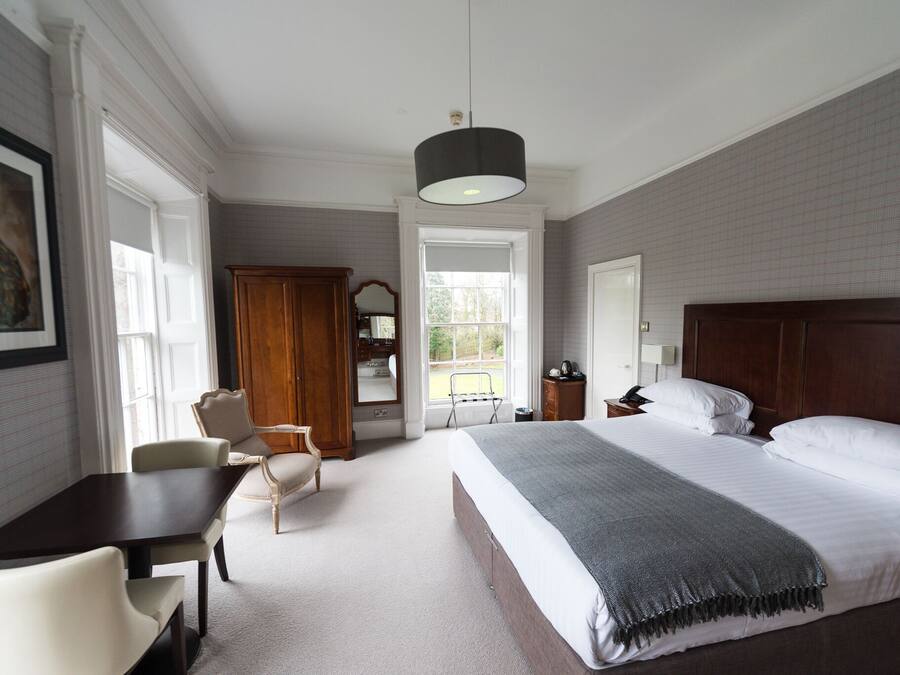 A bedroom with a large bed, a desk, and a chair. There is a window, a door, and a hanging light fixture.