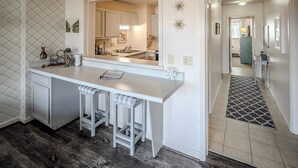 A kitchen with a white countertop, two bar stools, and a large mirror. There is a patterned wall and a rug leading to another room. - Sugar Ski Comfort Two Bedroom - Unit 625 (Sugar Mountain)