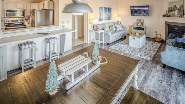 A modern kitchen with stainless steel appliances, a dining area with a wooden table and benches, a living room with a sofa and a TV, and a fireplace. - Sugar Ski Comfort Two Bedroom - Unit 625 (Sugar Mountain)