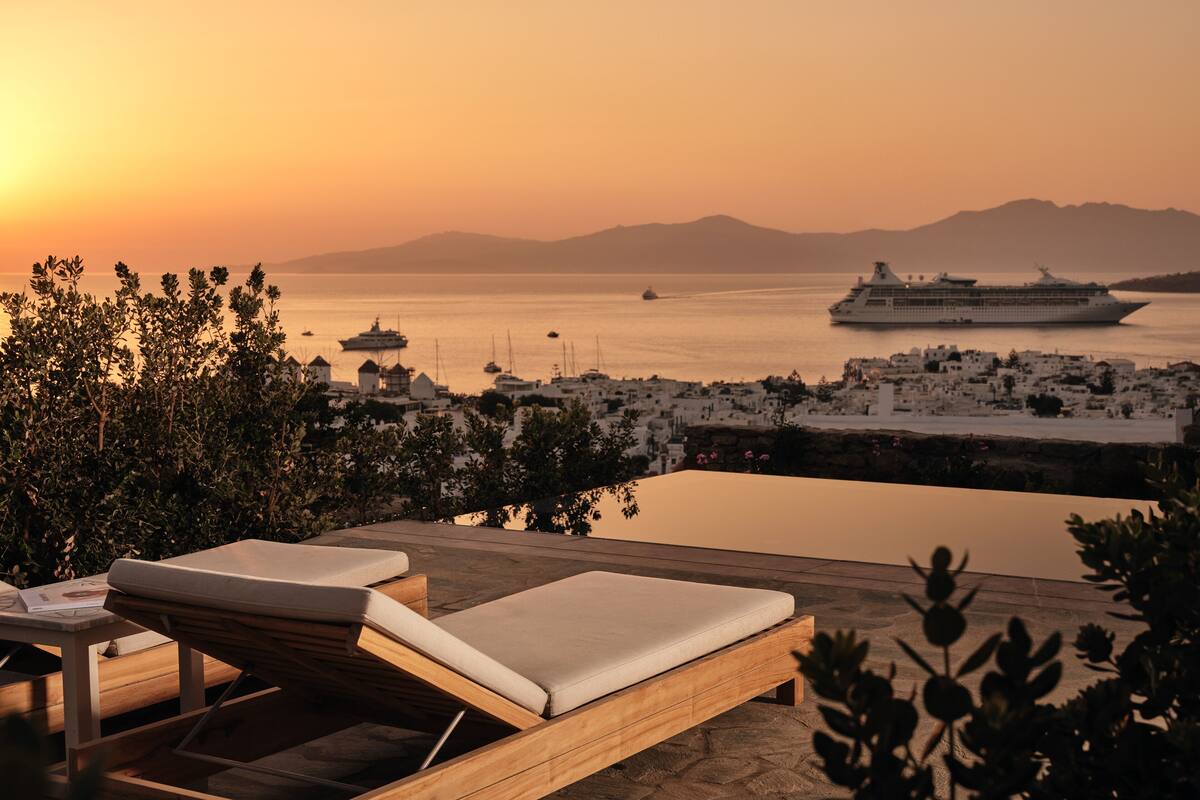 Un fauteuil de jardin en bois avec un coussin, une terrasse dallée de pierres et une vue sur une ville côtière, avec des bateaux et un navire de croisière visibles au loin.