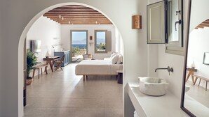 Un dormitorio moderno con una cama grande, un baño con lavamanos de mármol y vista al mar a través de un arco.