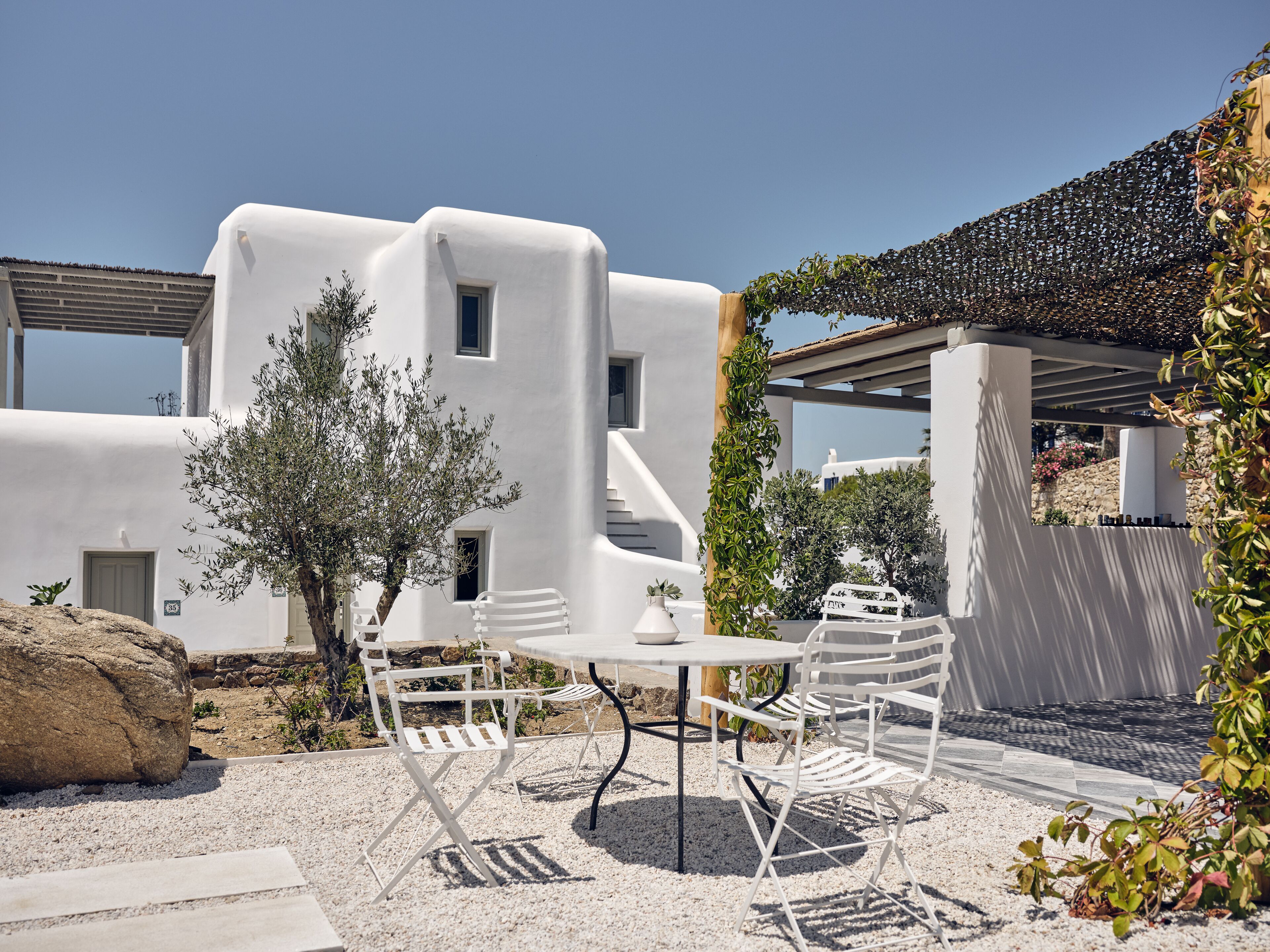 A white building with a stone wall and a pergola. There is a table and chairs in the foreground.