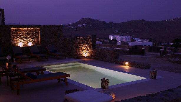 Une piscine à débordement aux murs de pierre, agrémentée de chaises longues et d’une table, le tout entouré d’un paysage montagneux.