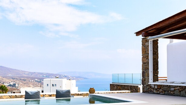 A pool area with a view of the sea and hills, stone walls, and a white building. - Ortus Light Blue Mykonian Luxury Villa (Mykonos)