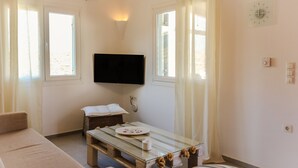 A room with a sofa, a glass-top table with a plate and a cup, a flat-screen TV mounted on the wall, and a window with curtains. - Ortus Light Blue Mykonian Luxury Villa (Mykonos)