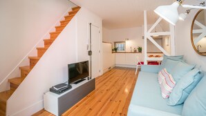 A modern living room with a wooden staircase, a flat-screen TV, a sofa with patterned cushions, and a dining area in the background.