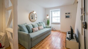 A cozy living room with a light green sofa, a round mirror, a small table with a TV, and wooden flooring.