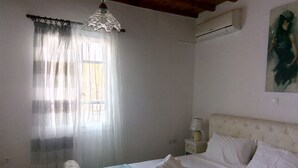 A bedroom with a bed, a nightstand, a lamp, a window with sheer curtains, and an air conditioning unit.