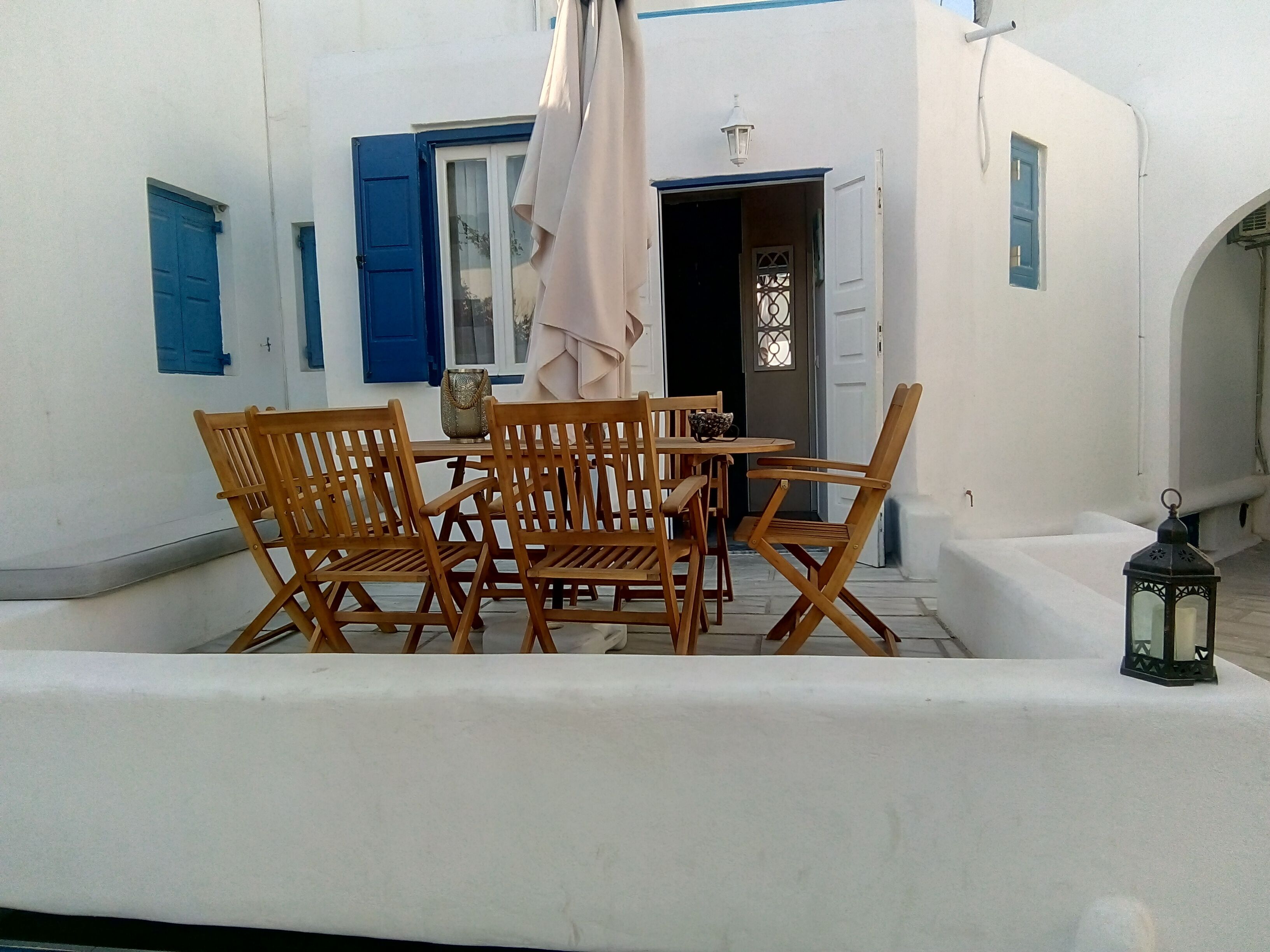 A white-walled building with blue shutters, a wooden table with chairs, and a lantern.