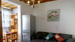 A compact living space with a wooden dining table, a sofa with patterned cushions, a refrigerator, and a chandelier.