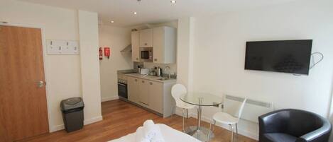 A hotel room with a bed, a black leather chair, a small kitchen area, a glass table, and a TV mounted on the wall.