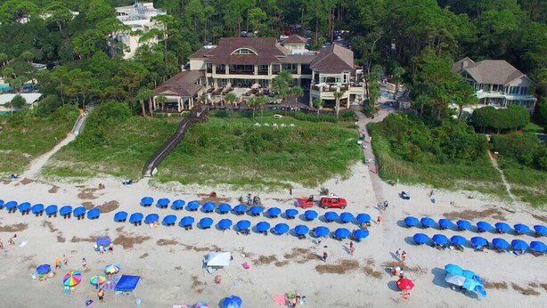Une vue aérienne d’une plage où l’on aperçoit de nombreux parasols bleus et une grande maison avec une allée menant à l’entrée.