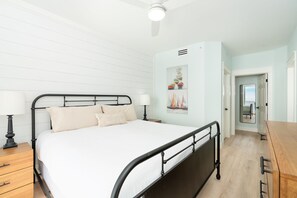 Une chambre à coucher comprenant un lit, des tables de chevet, un ventilateur de plafond et une porte donnant sur une autre pièce.