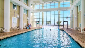 Una piscina interior con paredes de vidrio transparente, suelo de baldosas y varias sillas de descanso alrededor de la zona de la piscina.