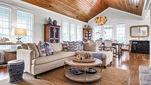 Un ampli saló amb un sostre fusta alt, un sofà seccional beix amb coixins estampats, una taula de centre rodona i una zona de menjador amb taula i cadires de fusta.