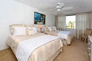 Two single beds with beige bedding and white pillows, a ceiling fan, and a painting adorn this serene hotel room with a view of palm trees.