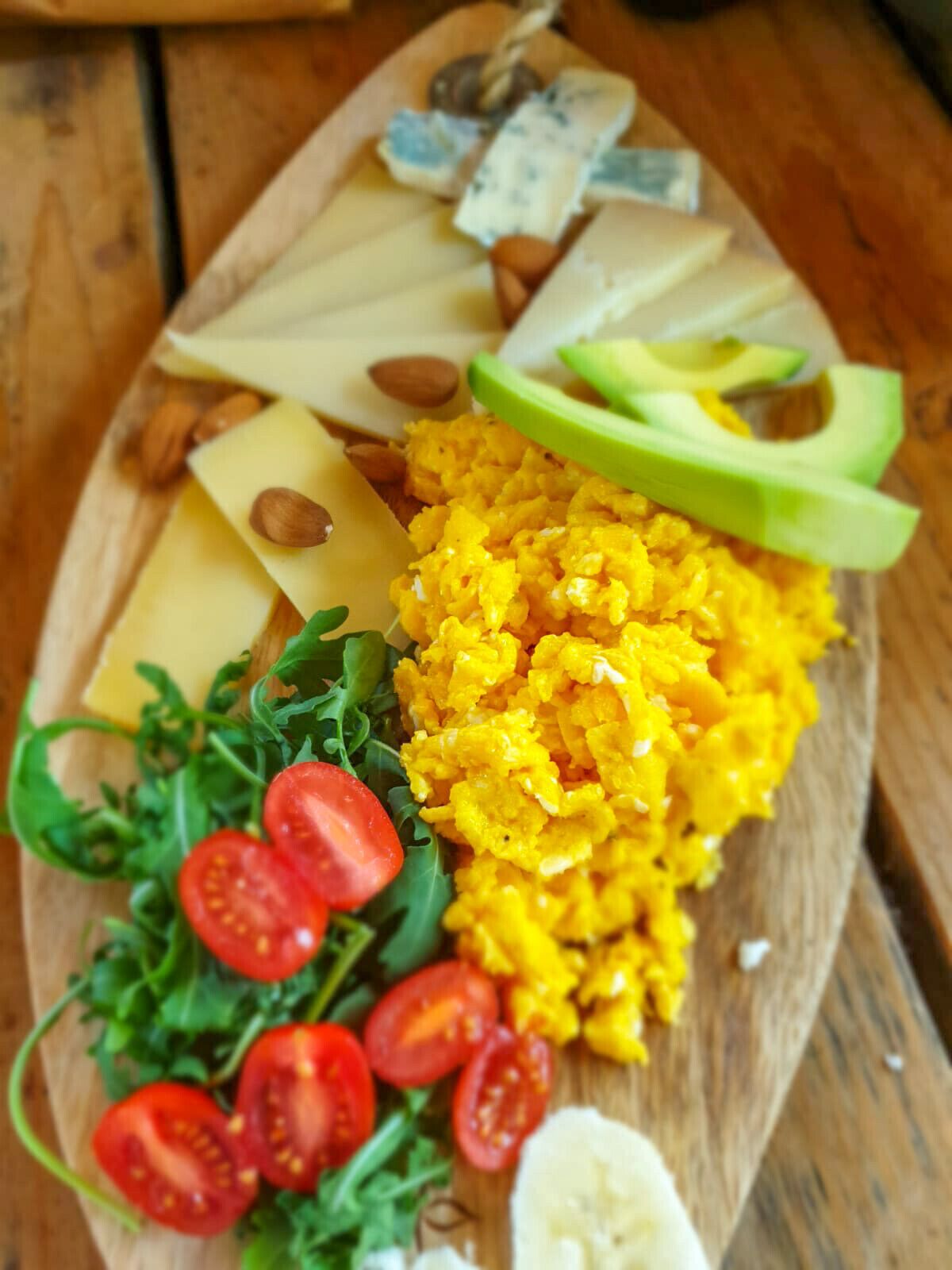 Een houten plank met diverse lekkernijen, waaronder kaas, amandelen, avocado en een salade met tomaten en bladgroenten.