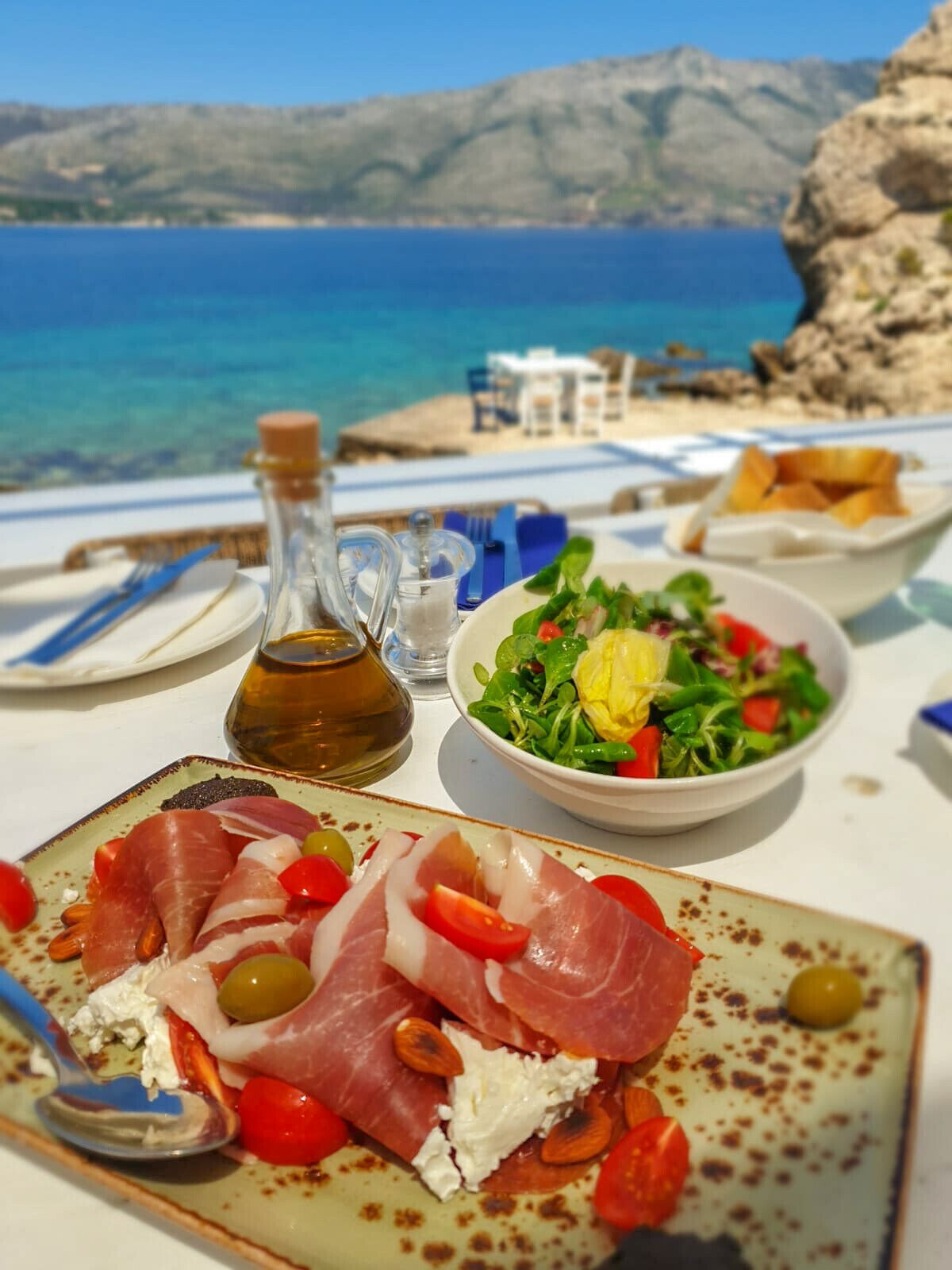 A table with a plate of prosciutto, olives, and tomatoes, a bowl of salad, and a bottle of olive oil.