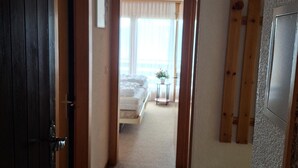 Ein Flur, der in ein Schlafzimmer mit Bett, kleinem Tisch und einem mit Vorhängen versehenen Fenster führt.