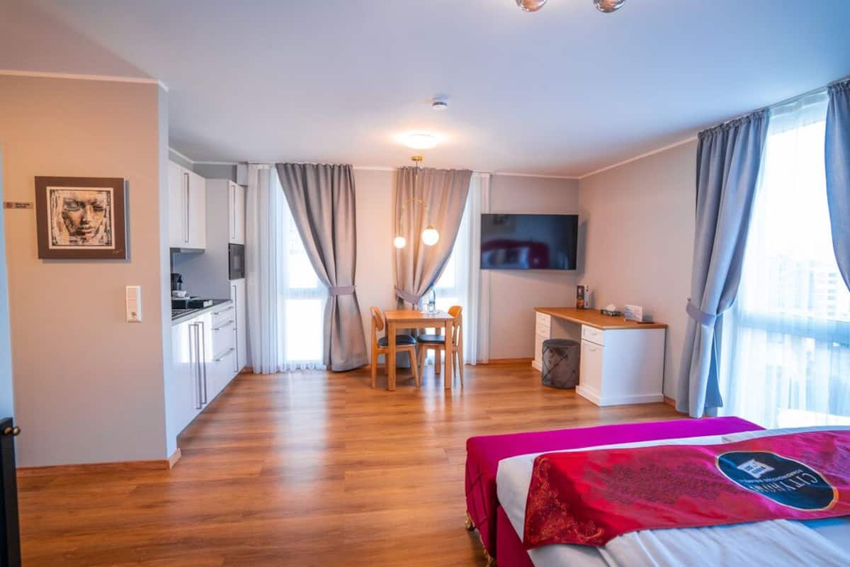 A modern hotel room with a wooden floor, a bed with a red and white bedspread, a desk with a chair, a television, and a kitchenette in the background.