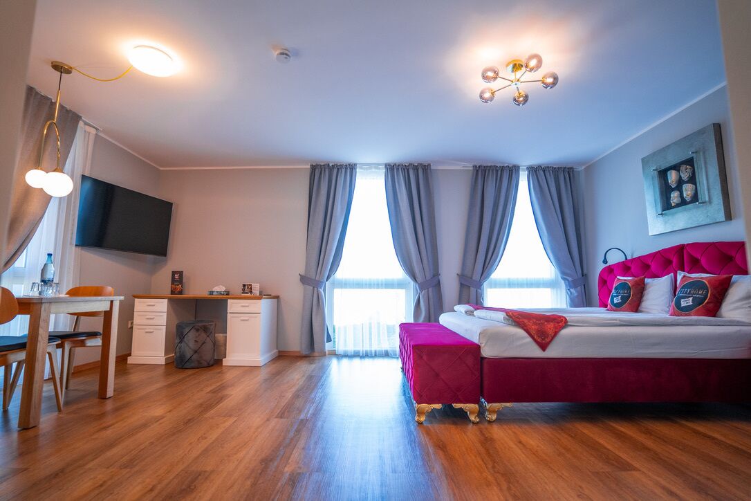 Una habitación de hotel moderna con piso de madera, un escritorio con silla, una cama con cabecera roja y un ventanal con cortinas.