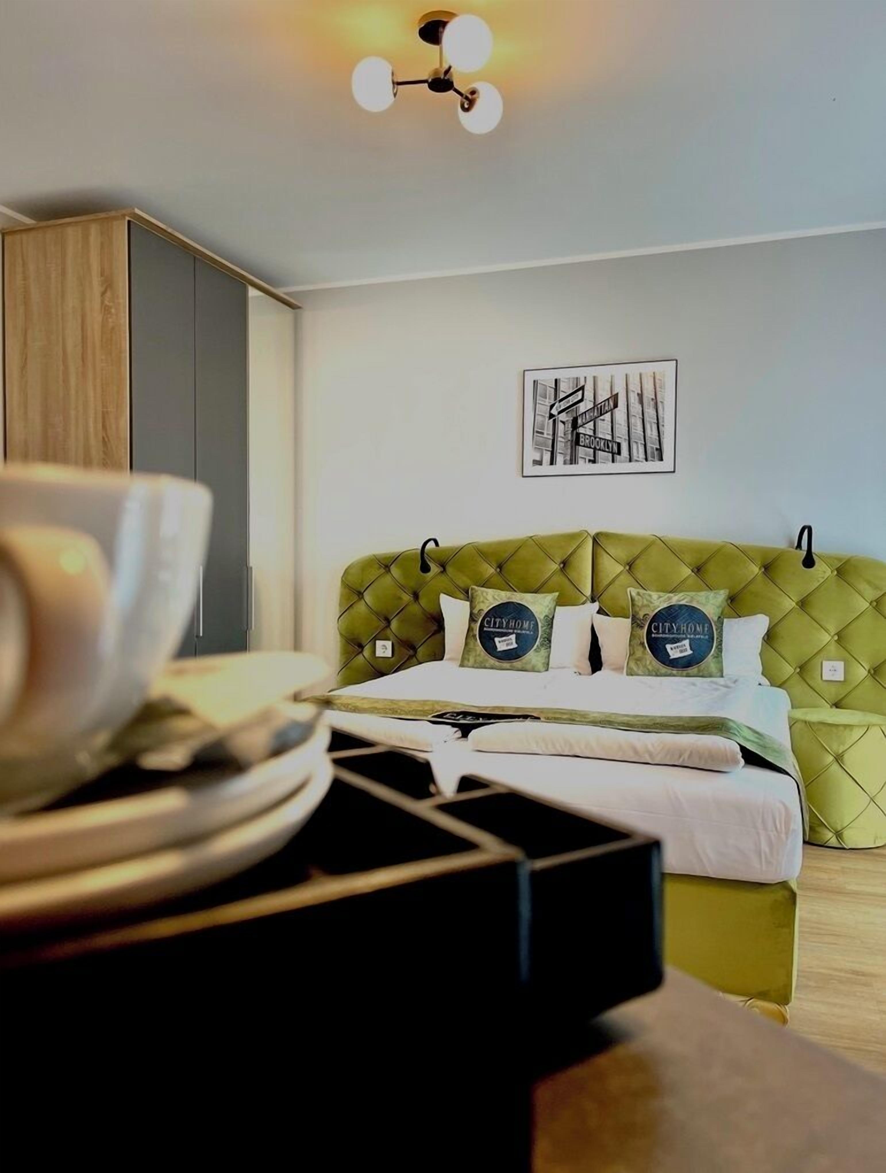 Una habitación de hotel con un cabecero verde tapizado, una cama con sábanas blancas, un cuadro enmarcado con una vista urbana y una lámpara de techo.