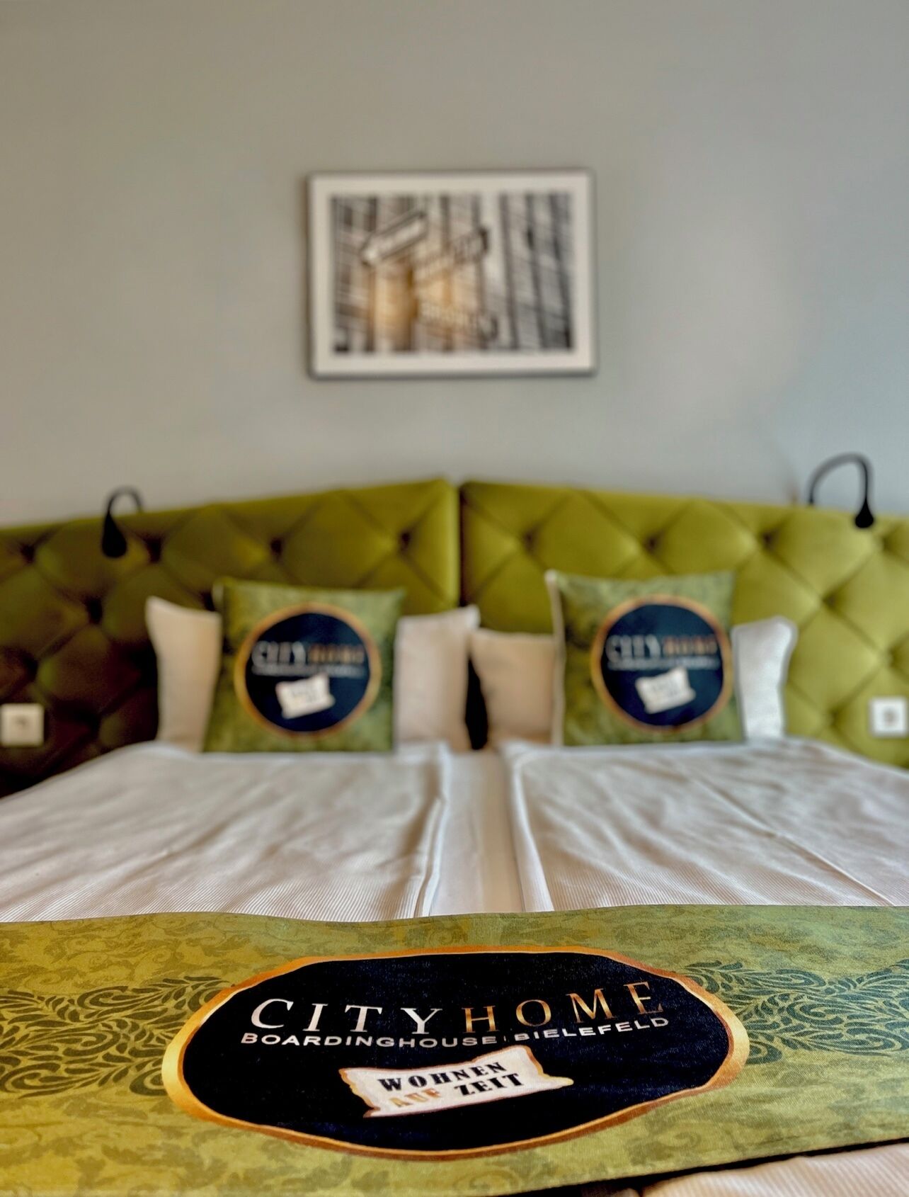 A bed with a green tufted headboard, two pillows with 'CITYHOME' branding, and a framed picture above.