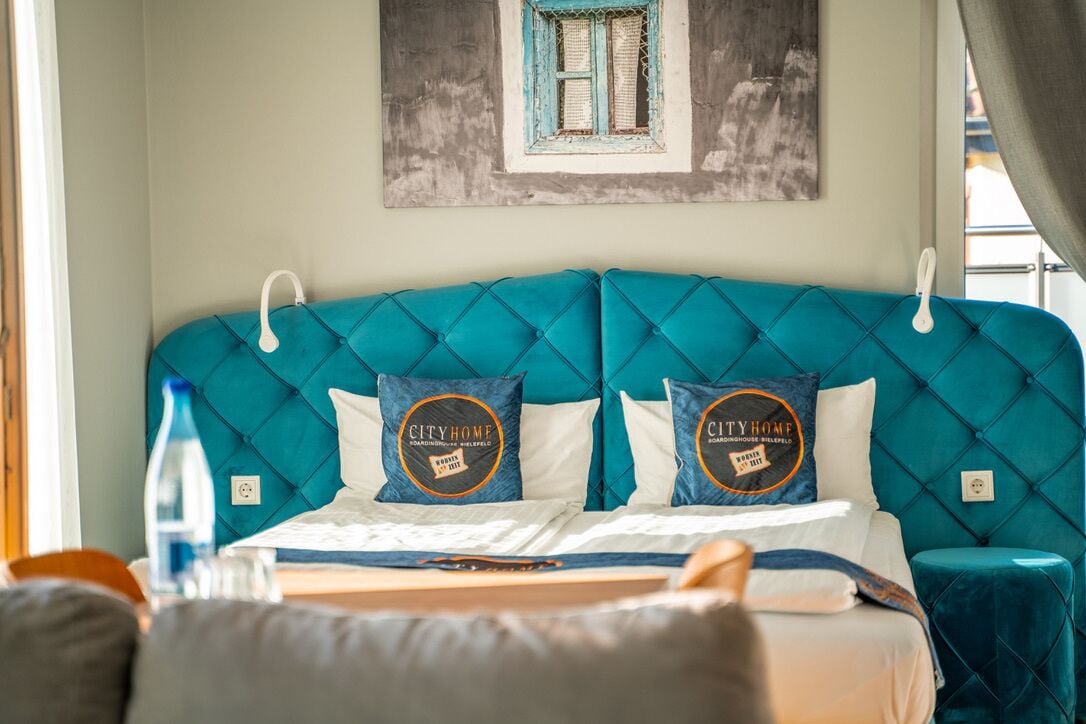 A hotel room with a teal upholstered headboard, two pillows with a circular logo, a bed with white linens, and a window with a view of a building.