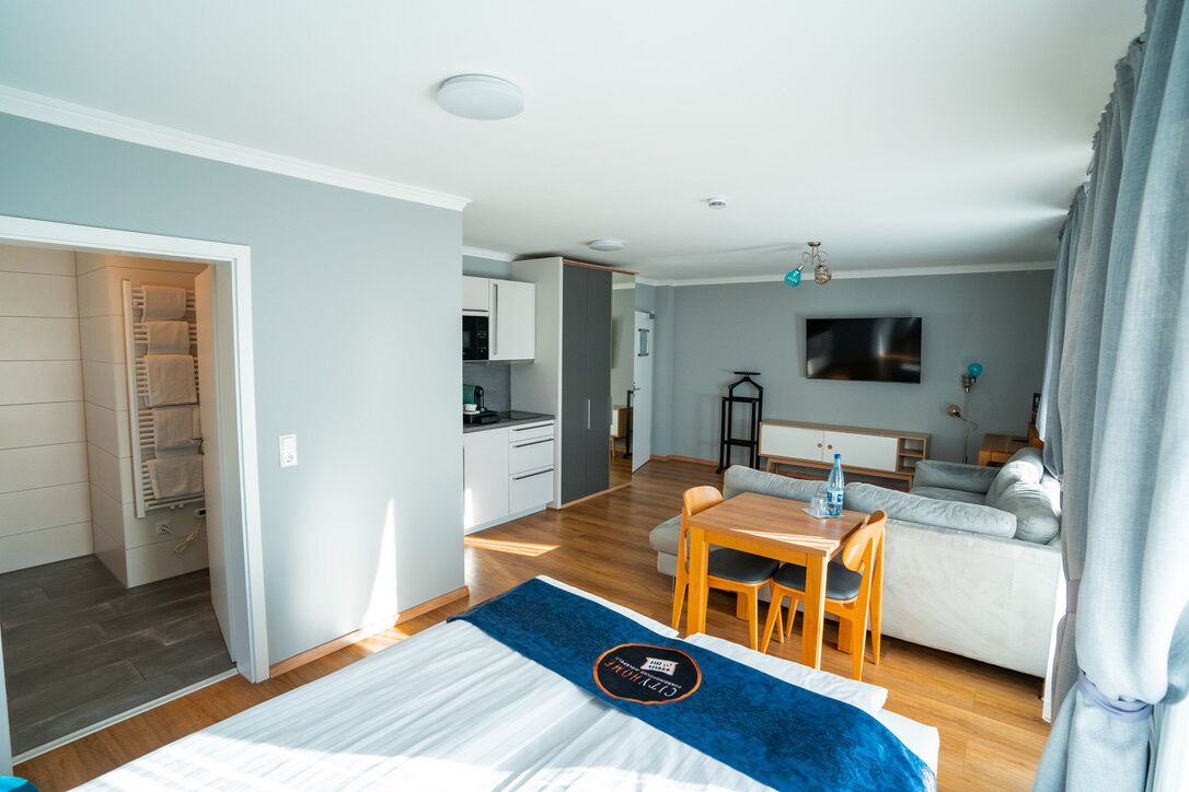 A modern hotel room with a bed, a dining area, a kitchenette, and a living area.