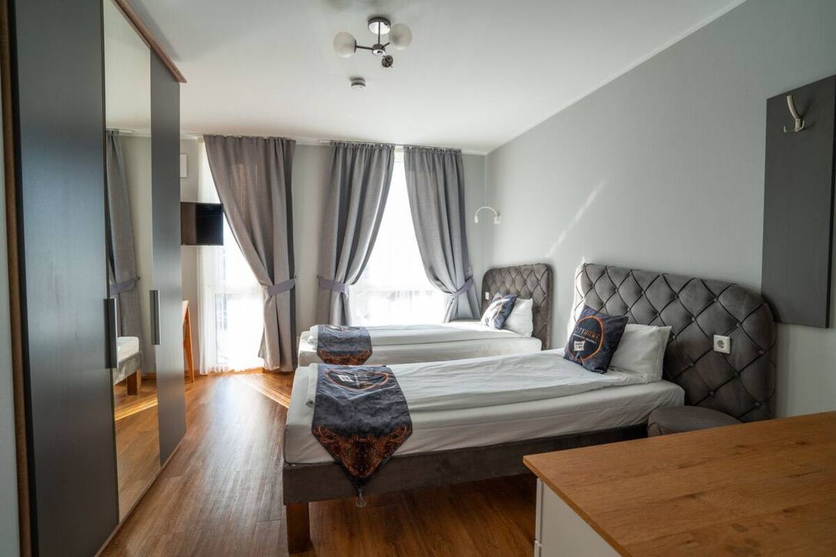A modern hotel room with two beds, a wooden floor, a ceiling fan, and a large window with curtains.