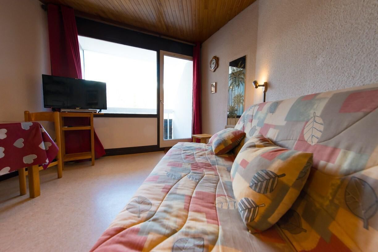 A cozy room with a bed, a TV on a stand, a small table, and a window with curtains.