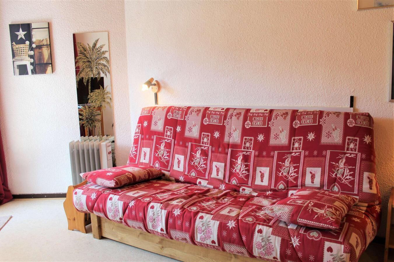 Et hyggeligt rum med en rød mønstret sofa, en lille træskammel, en radiator og en lampe på væggen.